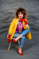 Wall Mural - A girl in a yellow jacket and blue jeans with an afro hairstyle sits on a chair. Fashion eighties, the era of disco. Studio photo on a gray background.
