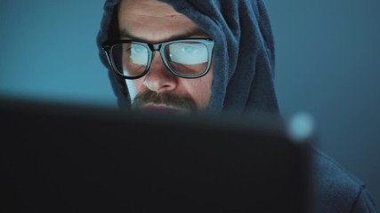 Male hacker in the hood and glasses working on a computer in a dark office room. Cybercrime concept