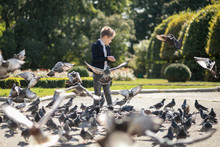Stylish Boy Of 7 Years In The Park Feeds Pigeons. There Are A Lot Of Birds. Baby Happy Feed Hungry Pigeons
