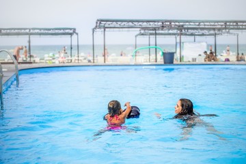 Wall Mural - Two little sisters girls are swimming in a large pool with clear blue water of mine near the hotel on the background of the sea and the beach. Concept vacation tropical hot country with children