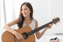 Carefree Pretty Asian Woman Is Practicing Acoustic Guitar. Happy Smiling Girl Enjoying With The Instrument In The White Room At Home.