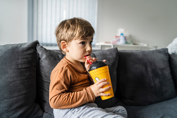 Portrait of small caucasian boy three or four years old sitting on the bed or sofa at home holding a plastic cup with straw drinking juice or soda at home alone in day