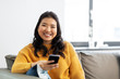 © Syda Productions - people, technology and leisure concept - happy smiling asian young woman in yellow sweater with smartphone sitting on sofa at home