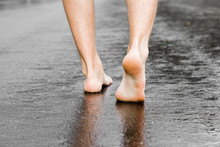 Young Man Barefoot Walking On Wet, Dark Black Asphalt After Warm Rain In Summer. Back View. Closeup.