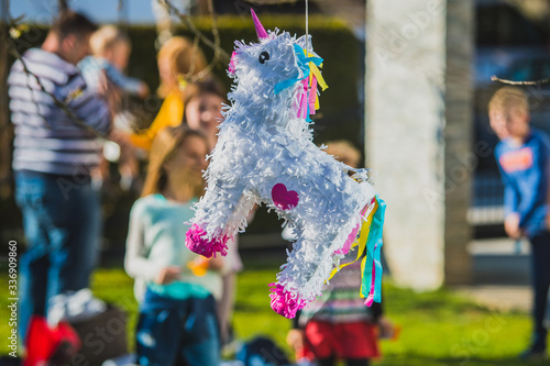 Young caucasian girl is hitting a white colored donkey shaped pinata hanging on the tree with kids cheering in the background. Festive activity during a birthday.