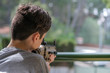 Teenager is playing with toy machine gun at home in quarantine on blurred sunny day background. The boy looks in gun sight from the balcony of apartment. Sad bored child. Weapon imitation.