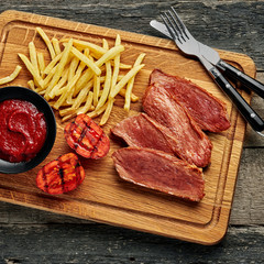 Wall Mural - Gourmet medium rare beefsteak with French fries and grilled cherry tomatoes on a cutting board with table knife and fork. Top view, directly above.