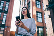 © BullRun - Below view of millennial blogger with cellular phone in hands looking away and pondering on ideas for content text, thoughtful woman in spectacles installing smartphone application outdoors