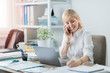 © Suteren Studio - Young businesswoman talking on smartphone and working on a laptop in a bright office. Woman preparing for a meeting with client to present project.