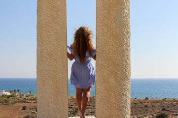 a beautiful young tourist on vacation in a light blue dress with long unpainted hair stands between two columns and looks at the sea and the blue sky