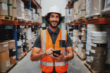 Young Smiling Male Worker In Warehouse Wearing Safety Helmet And Vest Standing Between Shelf With Goods Using Cellphone
