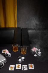 Staged photo of playing cards, casino chips, metal case and whiskey glasses on the black table against arm-chairs and a yellow curtain. The scene of the poker game is made in semi-darkness. 