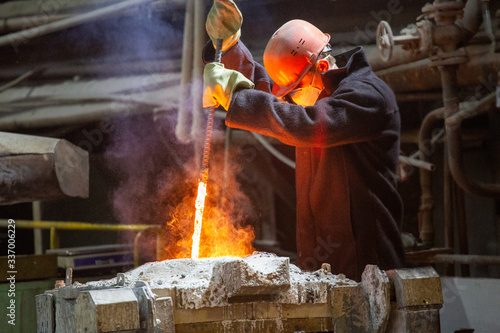 worker with hard hat and face mask stirs liquid metal in a furnace by steel bar