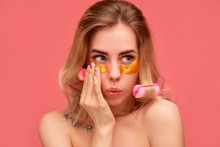 Close-up Girl With Rounded Lips, Looks Aside In Astonishment, Poses Blank Space. Young Woman With Curlers In Hair, Patches On Face (fabric Mask Under Eyes For Beauty), Isolated Pink Wall Studio