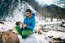Young Male Tourist Puts On Gloves Near A River In The Mountains..Beautiful Winter Landscape With Snow Covered Banks And Trees On Background. Climbing, Trekking, Active Life, Camping Concept.