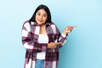 Wall Mural - Young latin woman woman isolated on blue background surprised and pointing side