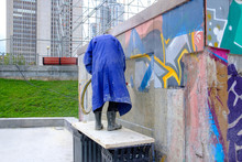 A Man In A Raincoat Washes Away Graffiti On The City Wall With A Stream Of Water And Chemicals