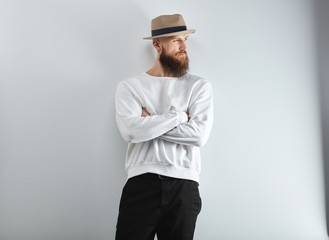 Wall Mural - Serious bearded man in hat standing against white wall arms crossed.