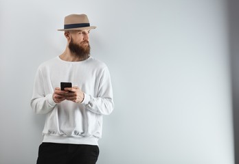 Wall Mural - Serious bearded man in hat standing against white wall using smart phone.