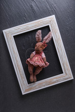 Toy Hare Made Of Coarse Fabric In A Light Frame On A Black Stone Background.