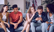 © Kostiantyn - Looking at the photos. Group of young happy multicultural people in casual wear looking at smartphone, enjoying time together while sitting on the sofa in the living room