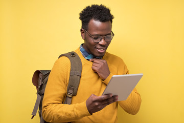 Canvas Print - Happy african american college student in glasses holding tablet on yellow wall. Studio shot