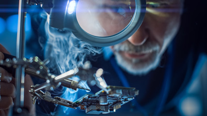 Fototapete - Electronics Maintenance and Repair Engineer Soldering Motherboard, Microchip and Circuit Board, Looking through Magnifying Glass, Consults Personal Computer. Close-up Low Angle Shot