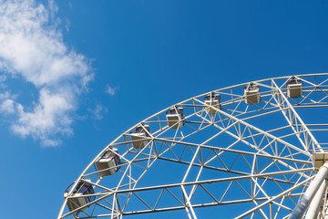 A part of white ferris wheel, cabins on background of blue sky and cloud in sunny summer day, bottom view.