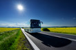 White bus driving on the asphalt road between the yellow flowering rapeseed fields under radiant sun in the rural landscape