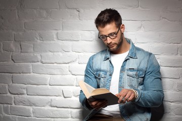 Poster - Smart casual man in glasses reading book, leading against brick wall. Copyspace.