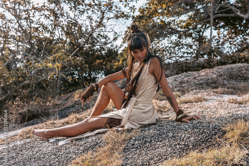 Plakat beautiful young stylish woman in elegant dress sitting on the rock at sunset