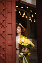 Young Attractive Woman In Hat, With Eyes Closed Posing Near Old Building With Antique Red Doors. In The Hands Woman Holding A Bouquet Of Yellow Tulips. Fashion And Lifestyle