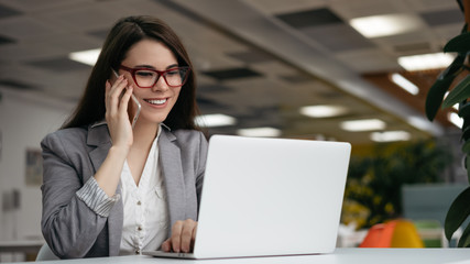 Successful businesswoman talking on mobile phone, working in office. Woman freelancer in stylish eyeglasses using laptop computer, looking at digital screen, communication at workplace 