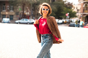 Fashionable young woman posing outside in a city street.Young beautiful stylish woman walking in brown jacket, blue jeans and red sweater, happy positive mood, sunny day.