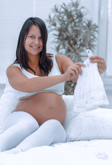 Wall Mural - pregnant woman considering children's shirt ,sitting on the bed.