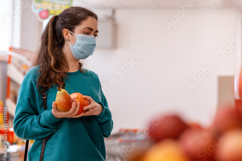 Photographie A young Caucasian woman with a medical mask on her face, holding a pear and a pomegranate, looking at a window with fruit