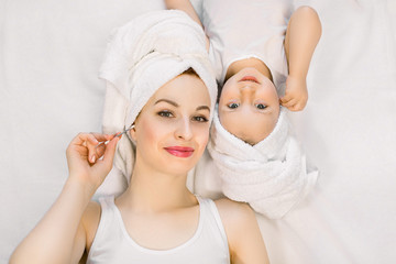 Wall Mural - Smiling mother and daughter in towels at bathtime. Young Caucasian woman mom with her little girl toddler, lying together on bed after bath or shower and cleaning their ears with cotton swab