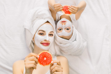 Wall Mural - Family beauty treatment in the bathroom. Pretty mother and daughter baby girl make a clay mask for face skin, lying on bed with towels on heads and holding slices of grapefruit in hands