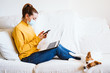 © Eva - young woman working on laptop and mobile phone, cute small dog besides. Sitting on the couch, wearing protective mask. Stay home concept during coronavirus covid-2019