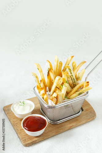 Yummy French Fries in a basket with tomato ketchup and mayonnaise in white bowls on a wooden board on a white background, garnished with herbs and cilantro. Wide Shot © Nehha