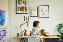 Female Artist At Her Workplace Working From Home. Young Woman Dressed In Jeans And Striped Shirt Sitting At The Table And Creating An Illustration.