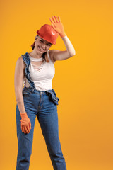 Builder girl in denim overalls and an orange helmet and gloves posing on a yellow background