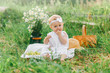 © Olga Gimaeva - little girl 1 year old sits in the grass in light clothes, eats  grapes, smiling, next to her is a basket and a bouquet of field daisies, childhood in the summer