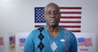 © vesperstock - Medium shot African American man in blue sweater and with silver cross, wears 'I Voted' sticker and poses proudly in front of polling booths with US flag and 'Vote' printed on them. Large US flag