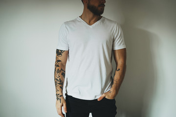 Wall Mural - Young athletic man with beard and tattoos wearing a blank white t-shirt and black jeans is standing on a white wall background. Horizontal mock up. Empty space for text o design.