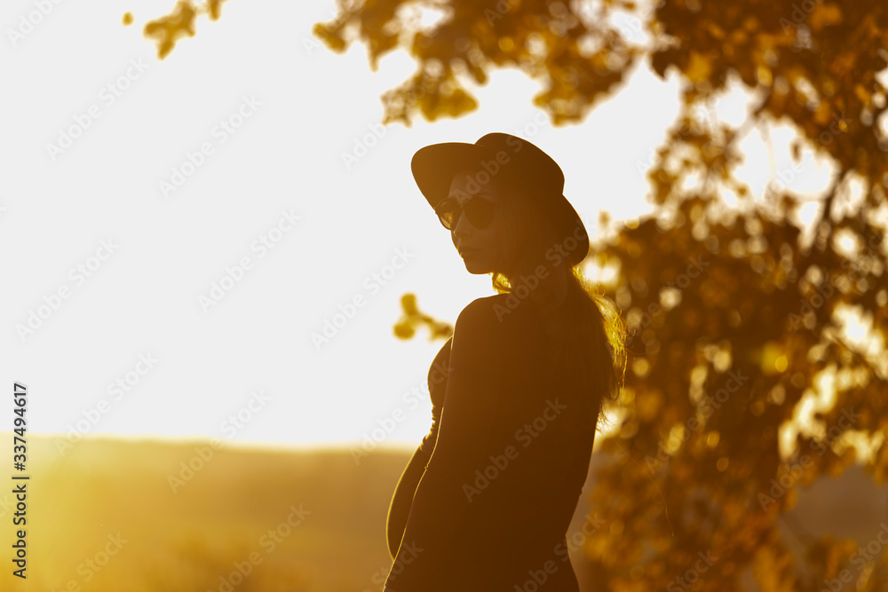 Beautiful pregnant woman in hat and sunglasses outside on warm sunny day. Facing to sun, looks at camera. Summer nature backsides. Yellow tones. Rest in park, meditation, retreat concept - obrazy, fototapety, plakaty 