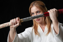 A Beautiful Girl With Blond Hair Holds Katana Sword With Two Hands In Front Of Her On A Black Background. Isolated Image