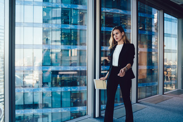 Wall Mural - Prosperous female proud ceo of corporation in formal wear passing glass panoramic window in office interior with modern tablet,serious intelligent business woman looking determinate on job with device