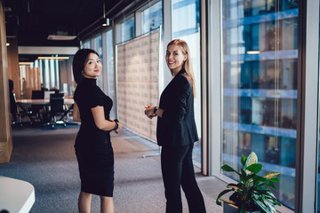 Wall Mural - Young women employees in formal wear standing in office satisfied with friendship on work talking to each other, smiling female employees enjoying successful career in corporation walking together