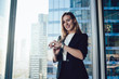© BullRun - Cheerful prosperous blonde woman in elegant outfit looking at camera standing near window in office,caucasian female manager checking time on trendy wearable wrist watch satisfied with bracelet